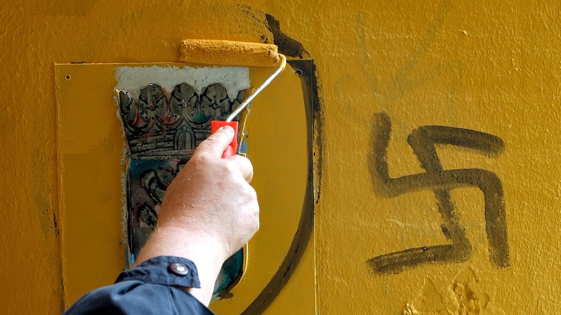 Ein Mann überstreicht Hakenkreuz-Schmierereien an der Außenfassade eines Gebäudes. Ein Mann überstreicht Hakenkreuz-Schmierereien an der Außenfassade eines Gebäudes.