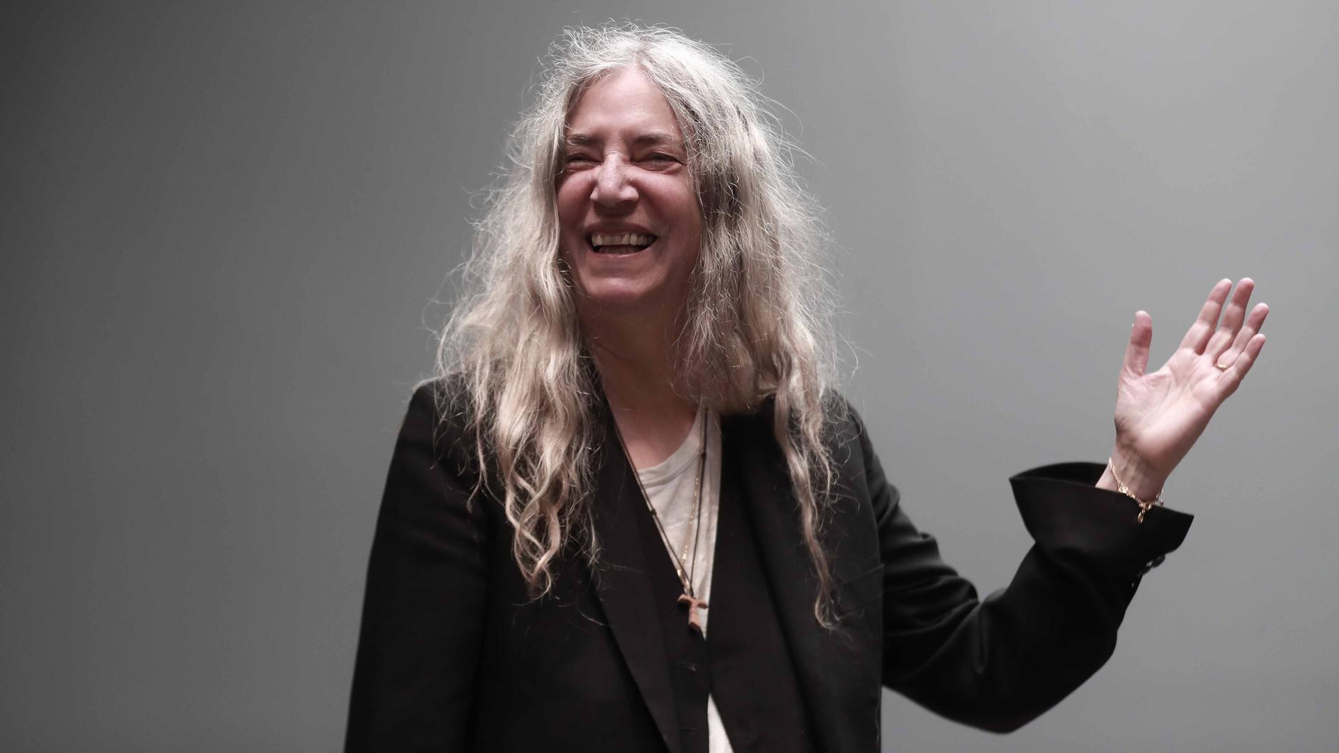 Patti Smith bei einem Fotoshooting in einem Studiosetting vor einem grauen Hintergrund. Sie trägt eine schwarze Blazer-artige Jacke, darunter ein cremefarbenes T-Shirt und eine lange Kette. Ihre langen, gewellten Haare fallen über ihre Schultern. Sie lacht und hat ihre linke Hand erhoben. Patti Smith bei einem Fotoshooting in einem Studiosetting vor einem grauen Hintergrund. Sie trägt eine schwarze Blazer-artige Jacke, darunter ein cremefarbenes T-Shirt und eine lange Kette. Ihre langen, gewellten Haare fallen über ihre Schultern. Sie lacht und hat ihre linke Hand erhoben.