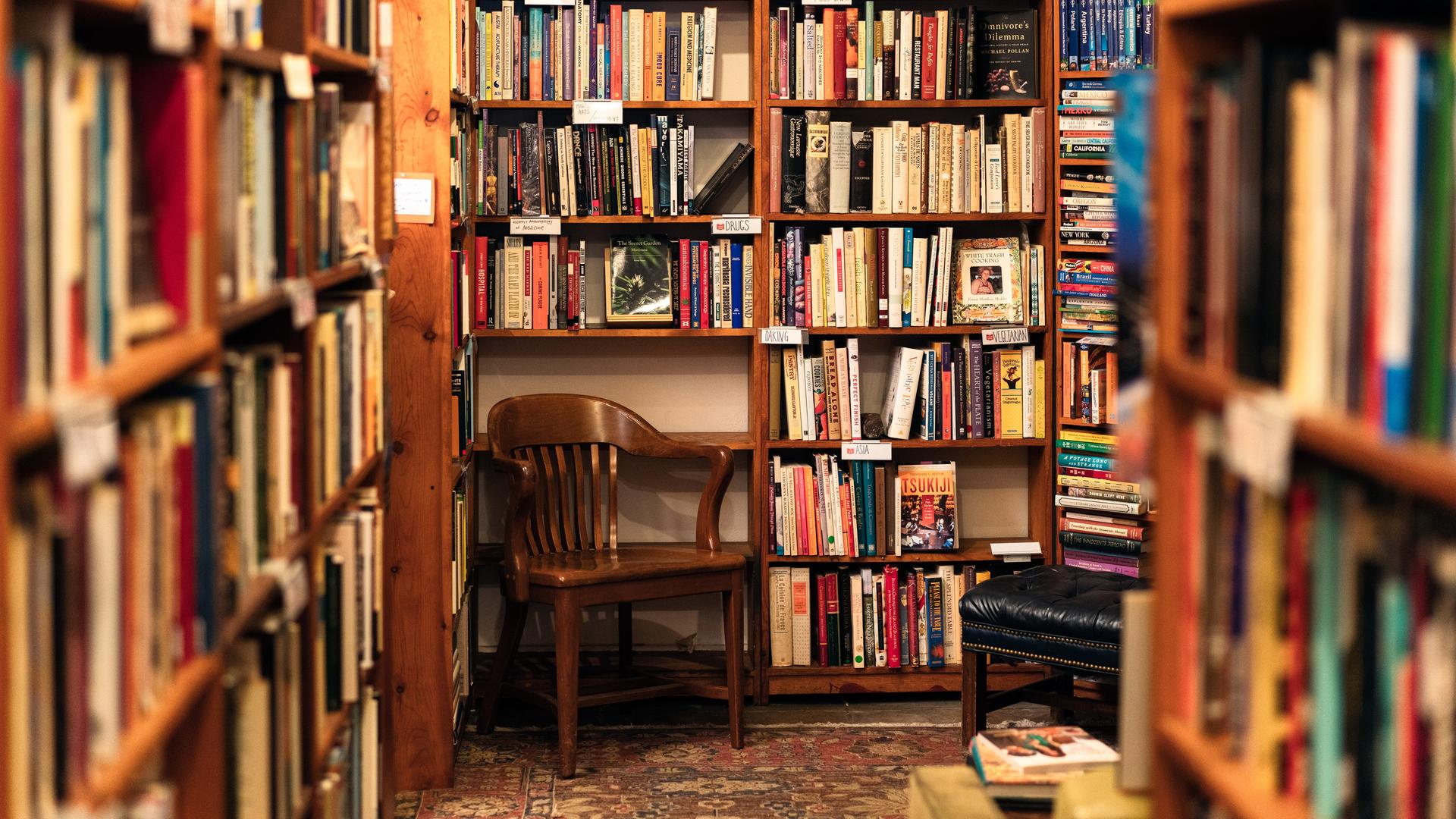 Ein hölzerner Stuhl steht in einer Ecke einer Secondhand-Buchhandlung mit zahlreichen Büchern auf hölzernen Regalen. Ein hölzerner Stuhl steht in einer Ecke einer Secondhand-Buchhandlung mit zahlreichen Büchern auf hölzernen Regalen.