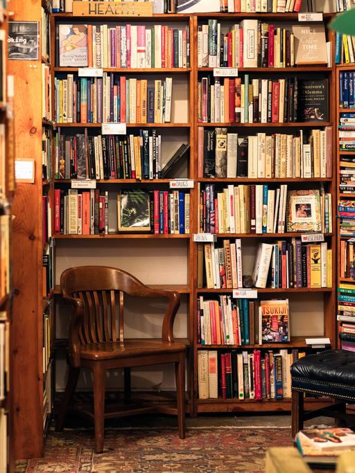 Ein hölzerner Stuhl steht in einer Ecke einer Secondhand-Buchhandlung mit zahlreichen Büchern auf hölzernen Regalen.