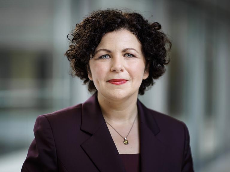 Amira Mohamed-Ali (Die Linke) trägt einen dunkel violetten Blazer und blickt aufmerksam in die Kamera.